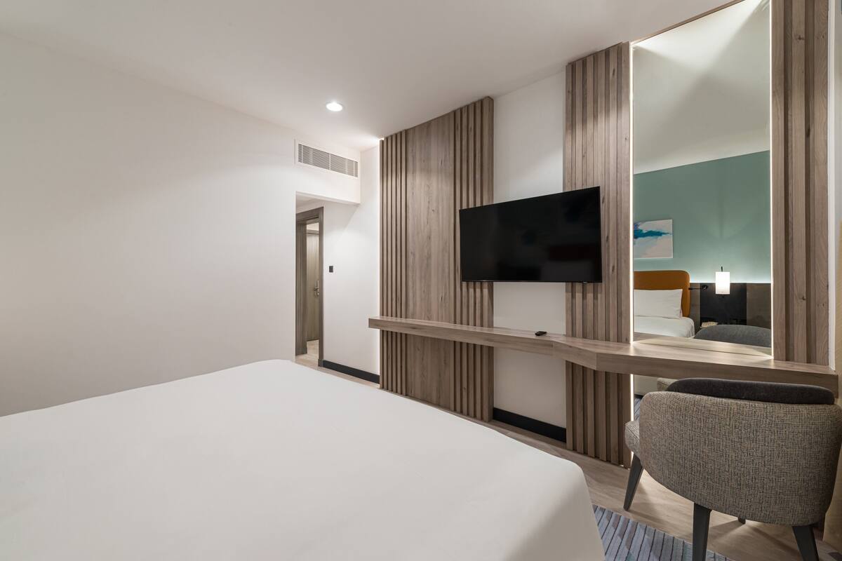 A modern hotel room with a large bed, a flat-screen TV mounted on a wooden panel wall, a mirror, and a seating area with a chair.