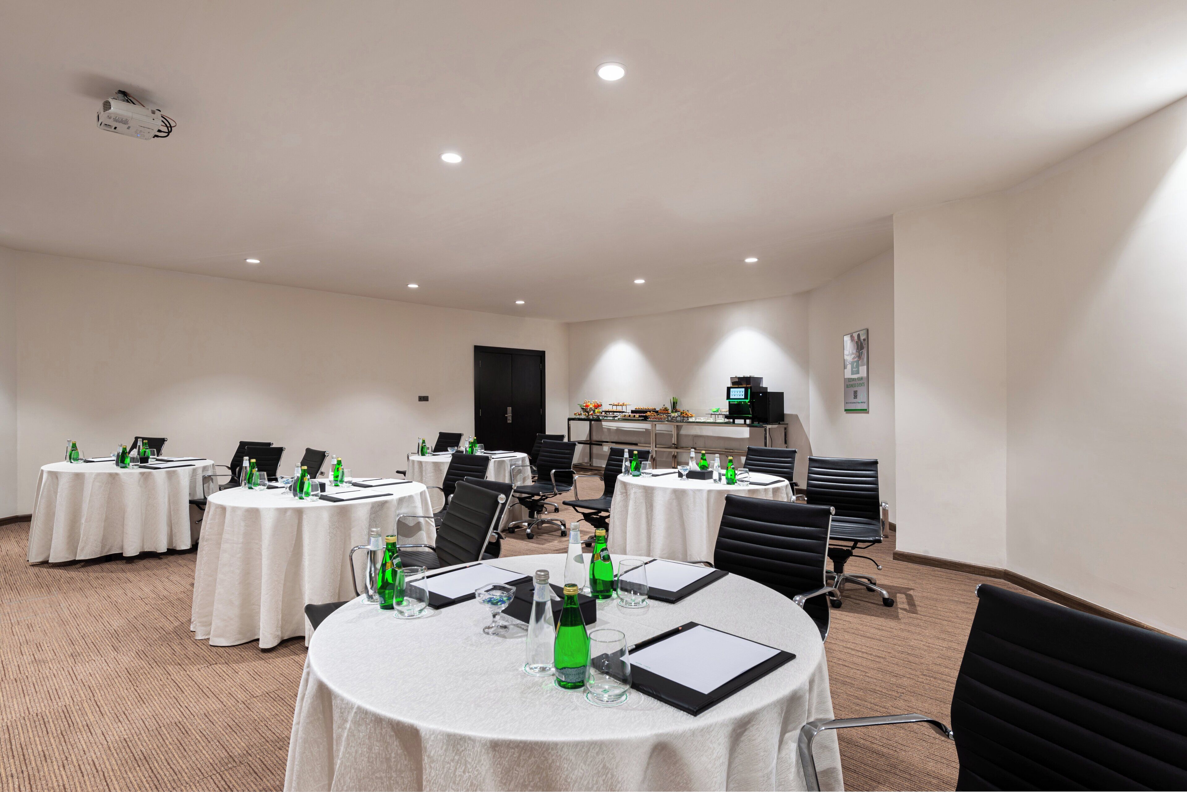 A conference room with multiple tables, chairs, and a projector. Tables are set with water bottles, glasses, and notepads.