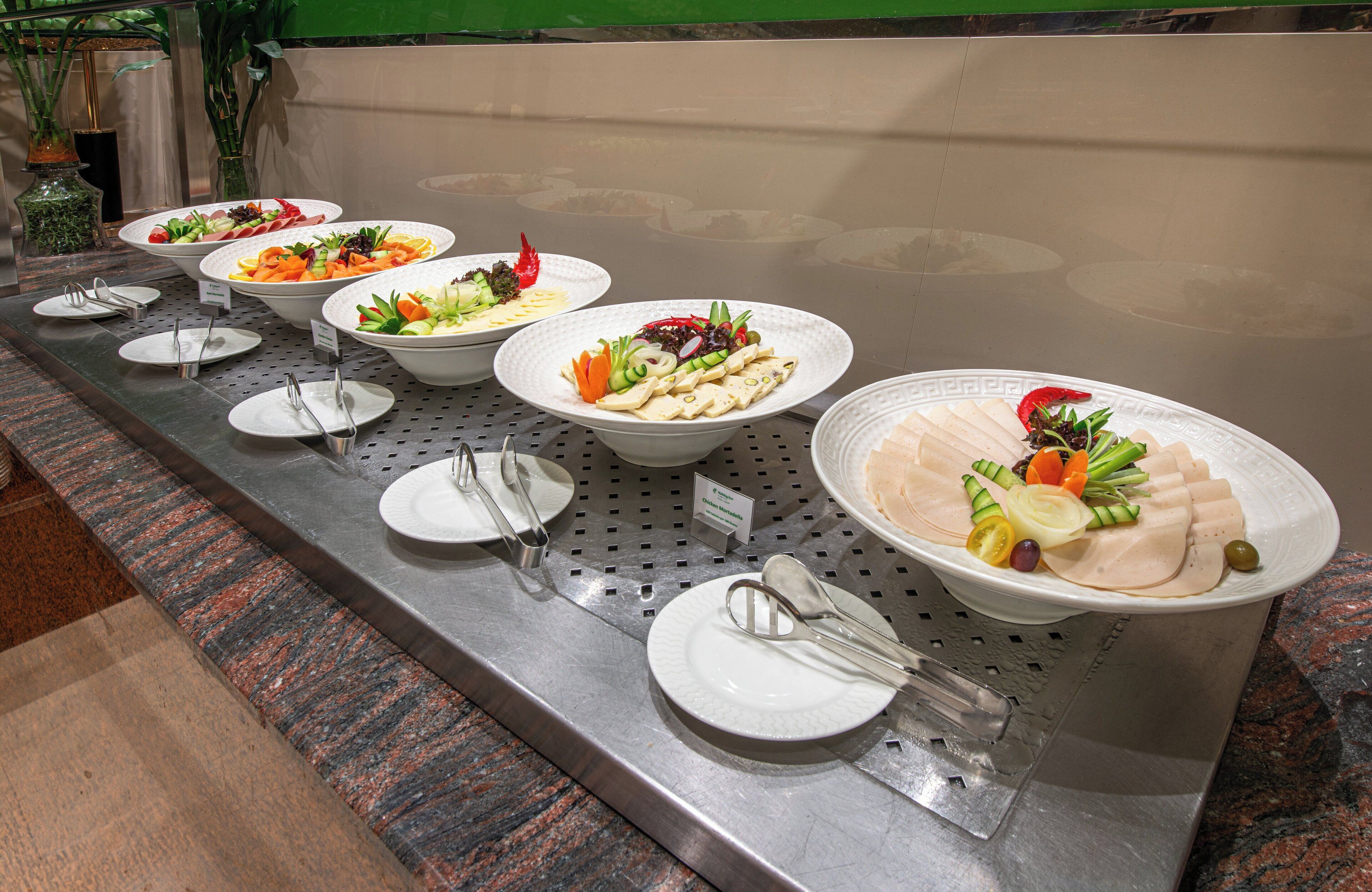 Un bufé con diversos platos, entre ellos ensaladas, frutas y carnes, dispuesto en un mostrador de metal con platos y cubiertos.
