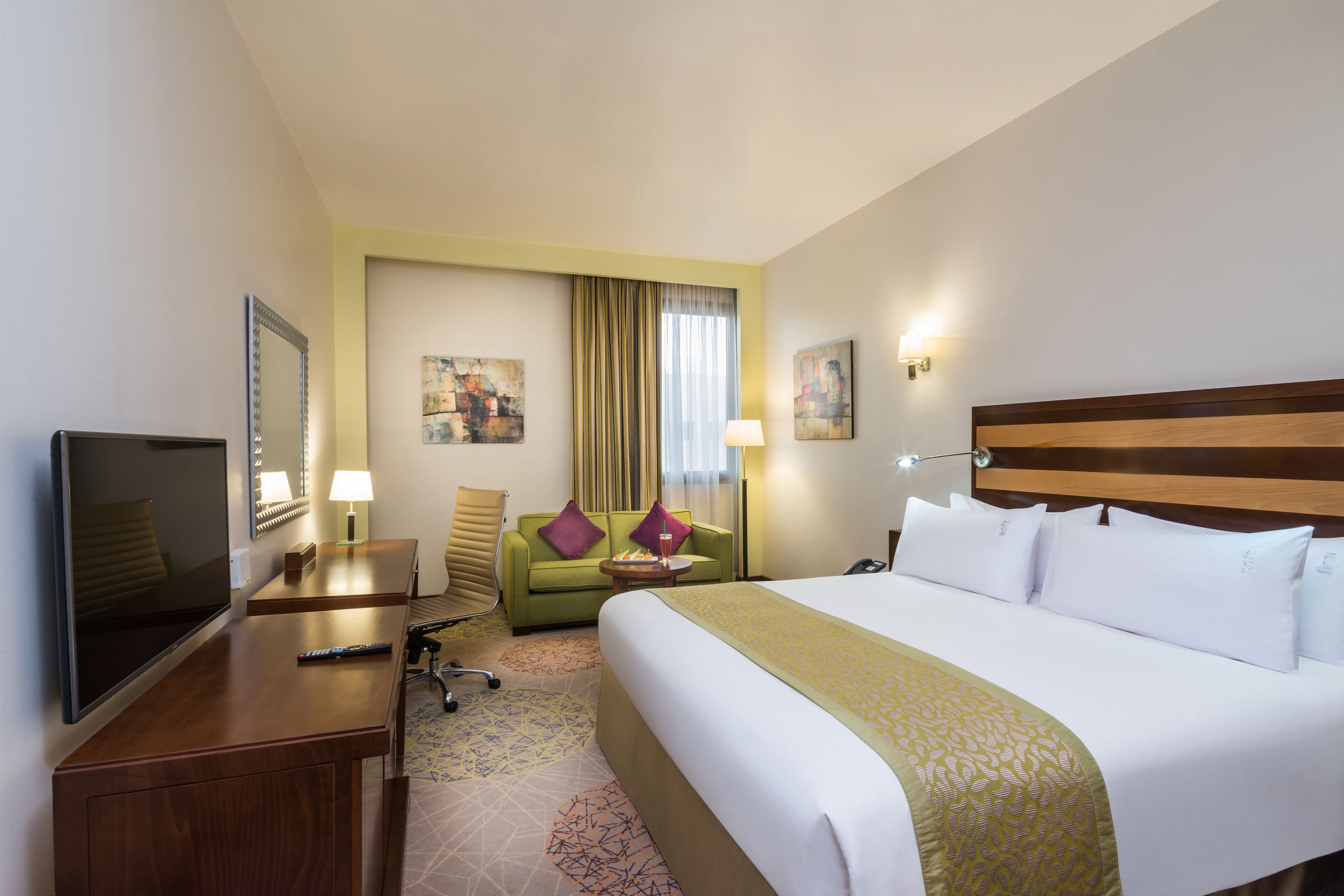 Una habitación de hotel con una cama grande, un escritorio con televisor, un sofá verde y dos cuadros en la pared.