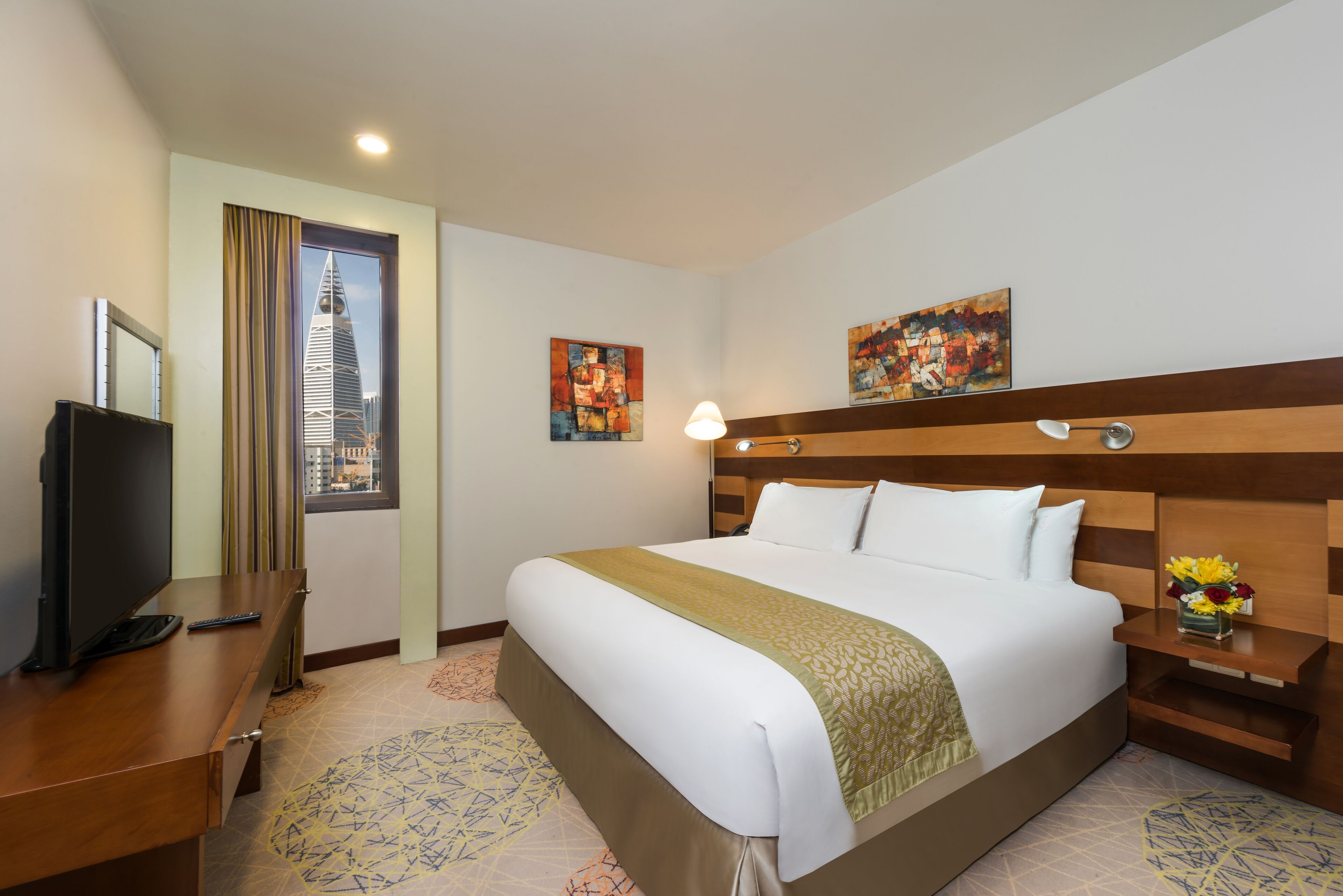A hotel room with a large bed, a desk with a TV, a nightstand with a lamp, and artwork on the walls.