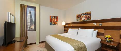 A hotel room with a large bed, a desk with a TV, a nightstand with a lamp, and artwork on the walls.