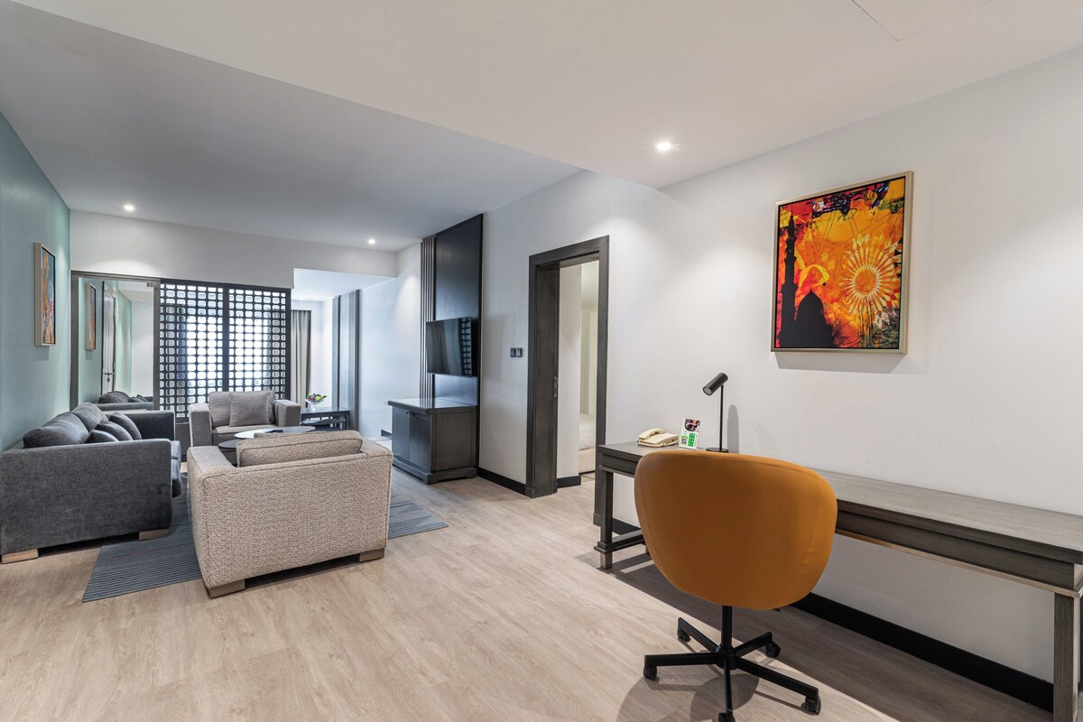 Een moderne woonkamer met een zithoek, een bureau met stoel, en een groot schilderij aan de muur.