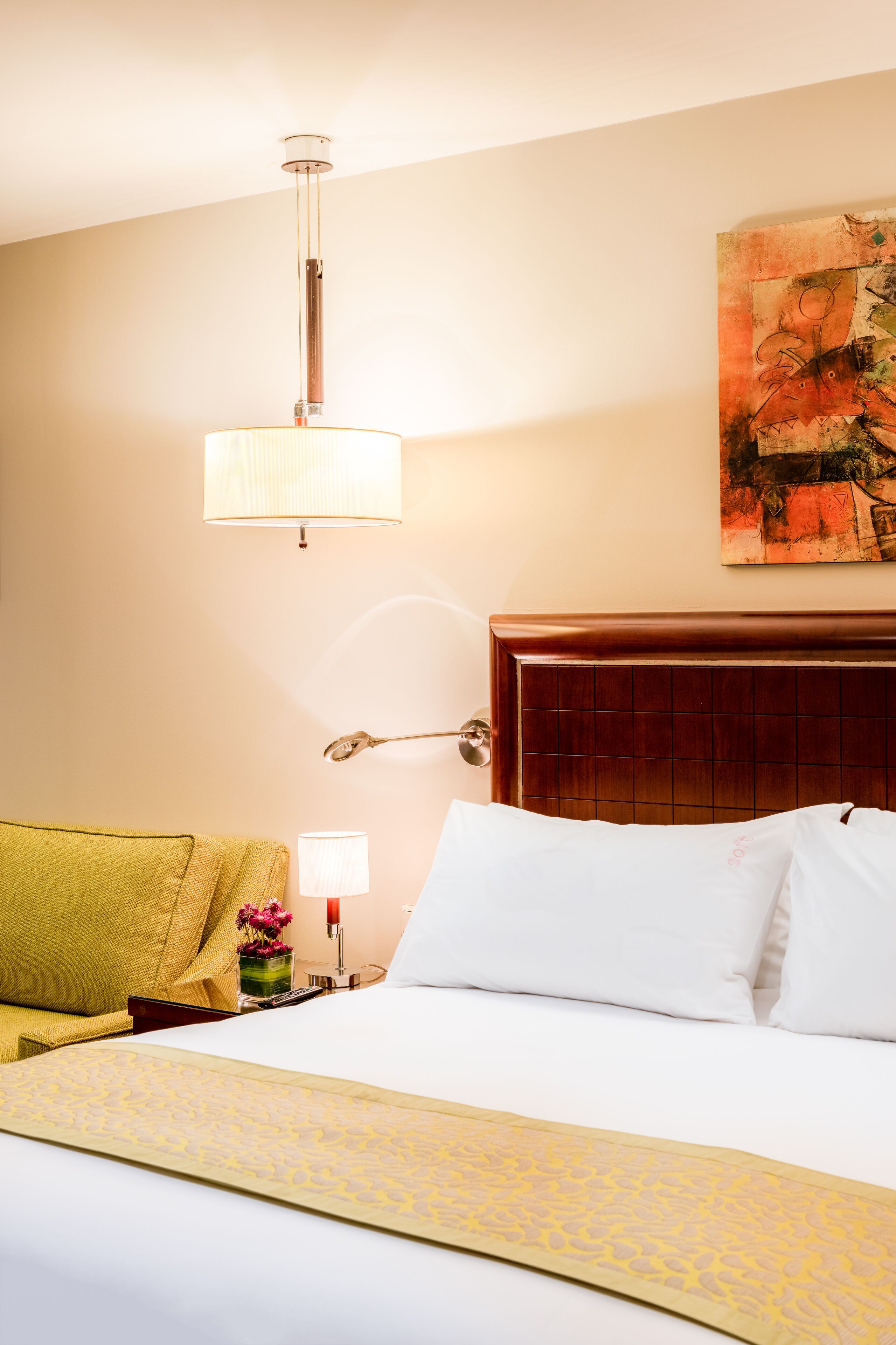 A hotel room with a bed, a bedside lamp, a nightstand, a vase with flowers, and a painting on the wall.