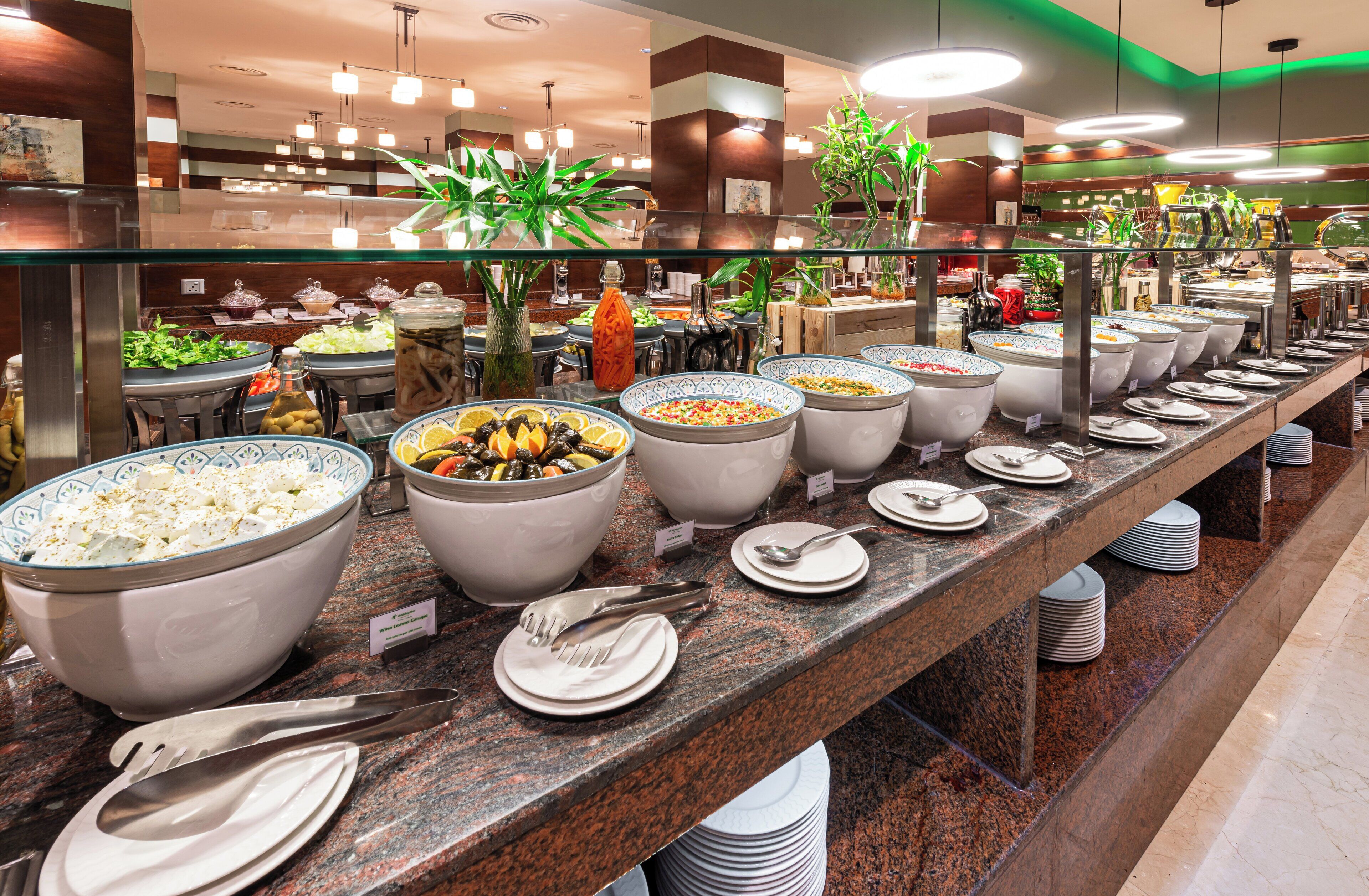 Un bufé con diversas opciones de comida, entre ellas ensaladas, frutas y otros platos, dispuesto en un mostrador largo con platos y cubiertos.