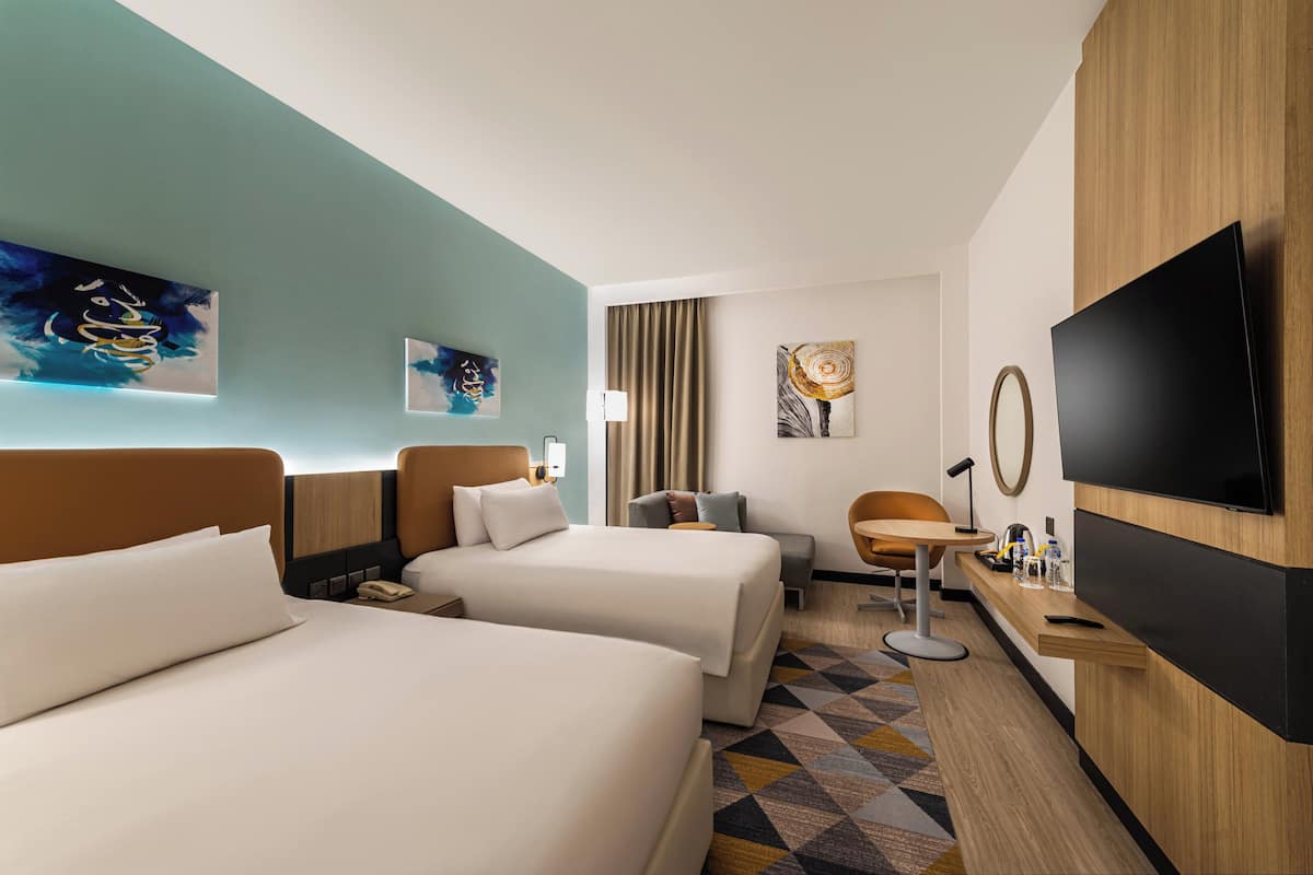 A hotel room with two beds, a desk, a TV, and artwork on the walls.