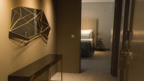 Un pasillo con un cuadro de arte geométrico en la pared, un largo banco y un acceso a un dormitorio con cama y mesita de noche.