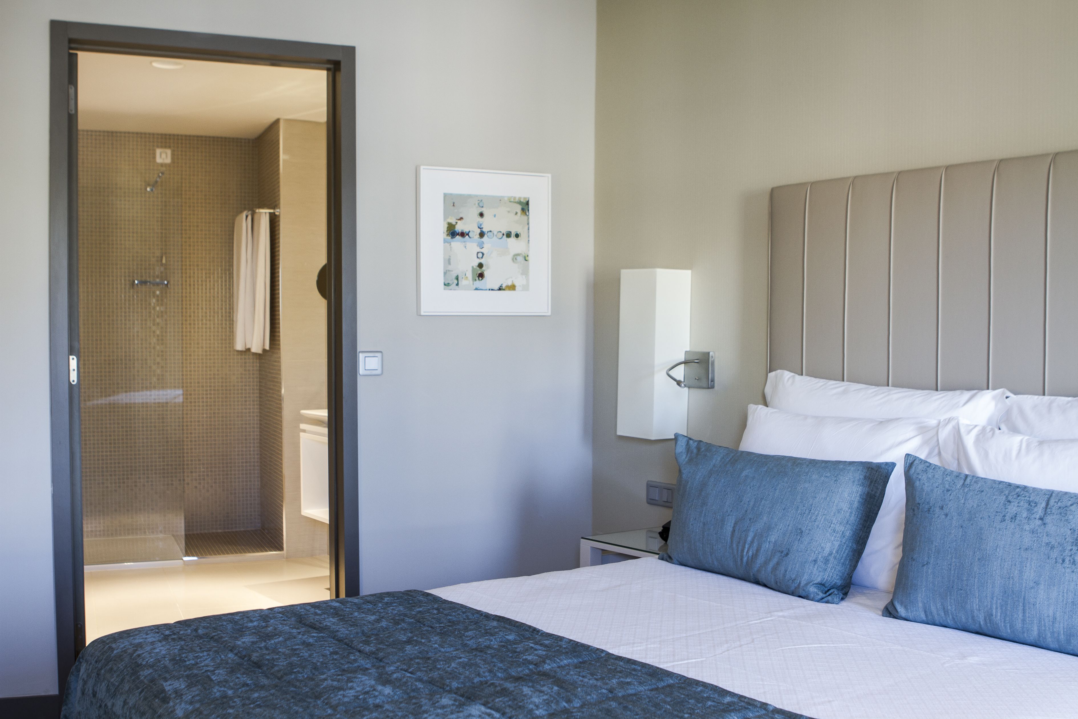 A hotel room with a bed, a nightstand, a wall-mounted lamp, a framed artwork, and a shower area.