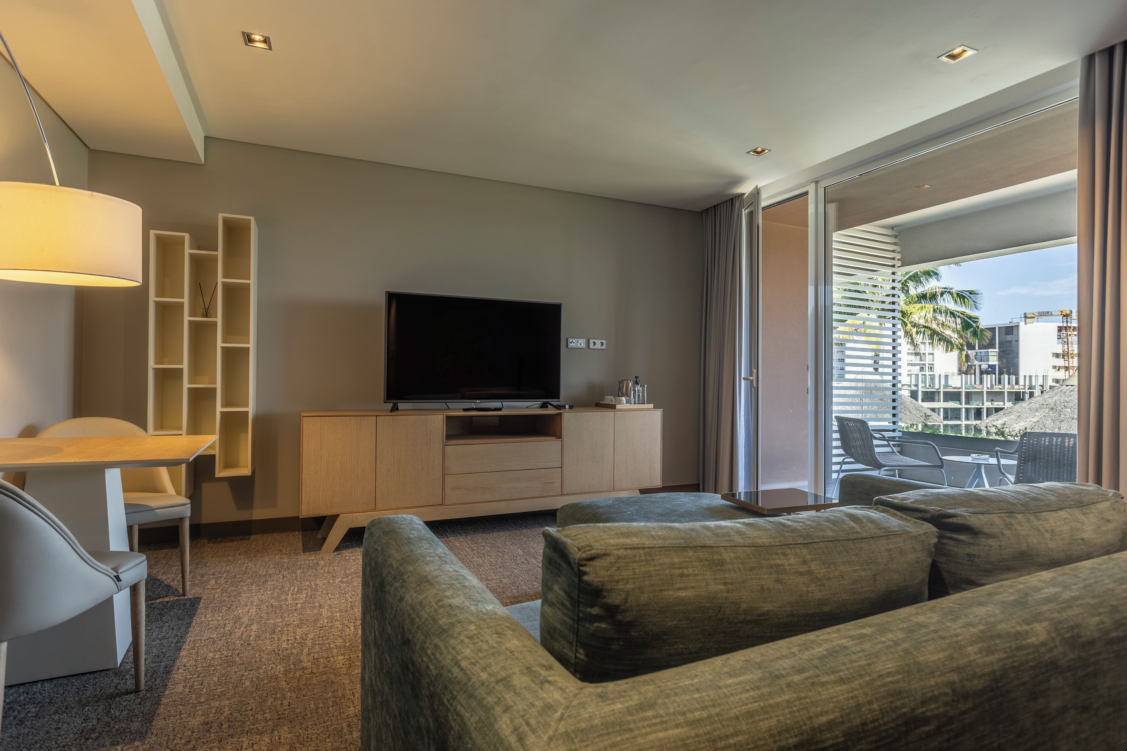 Una habitación de hotel moderna con una televisión de pantalla plana sobre un mueble de madera, un sofá, una mesa de comedor con sillas y vistas a la ciudad a través de amplios ventanales.