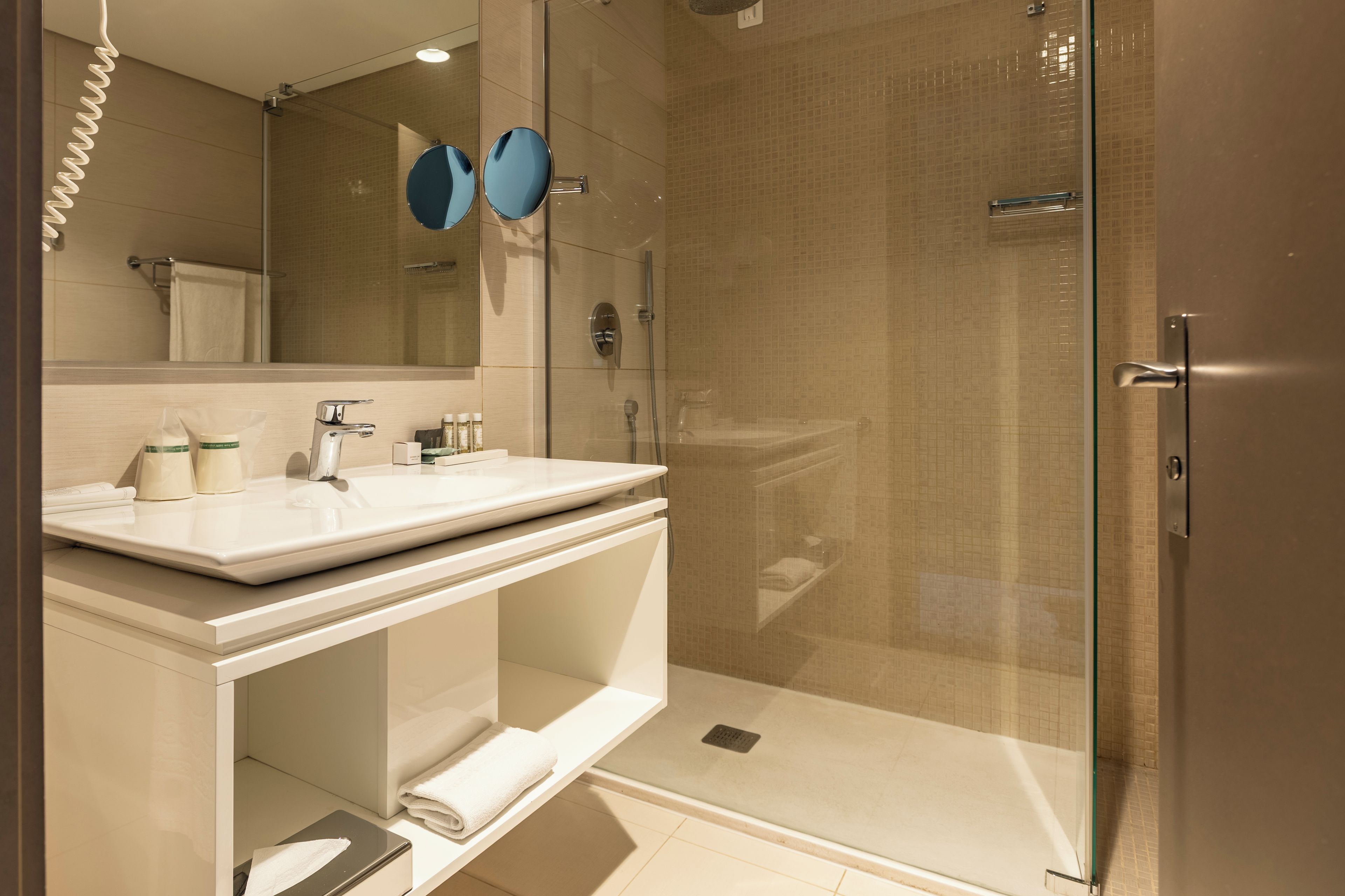 Un baño moderno con ducha envuelta en vidrio, un lavamanos blanco y un espejo con dos espejos redondos encima.
