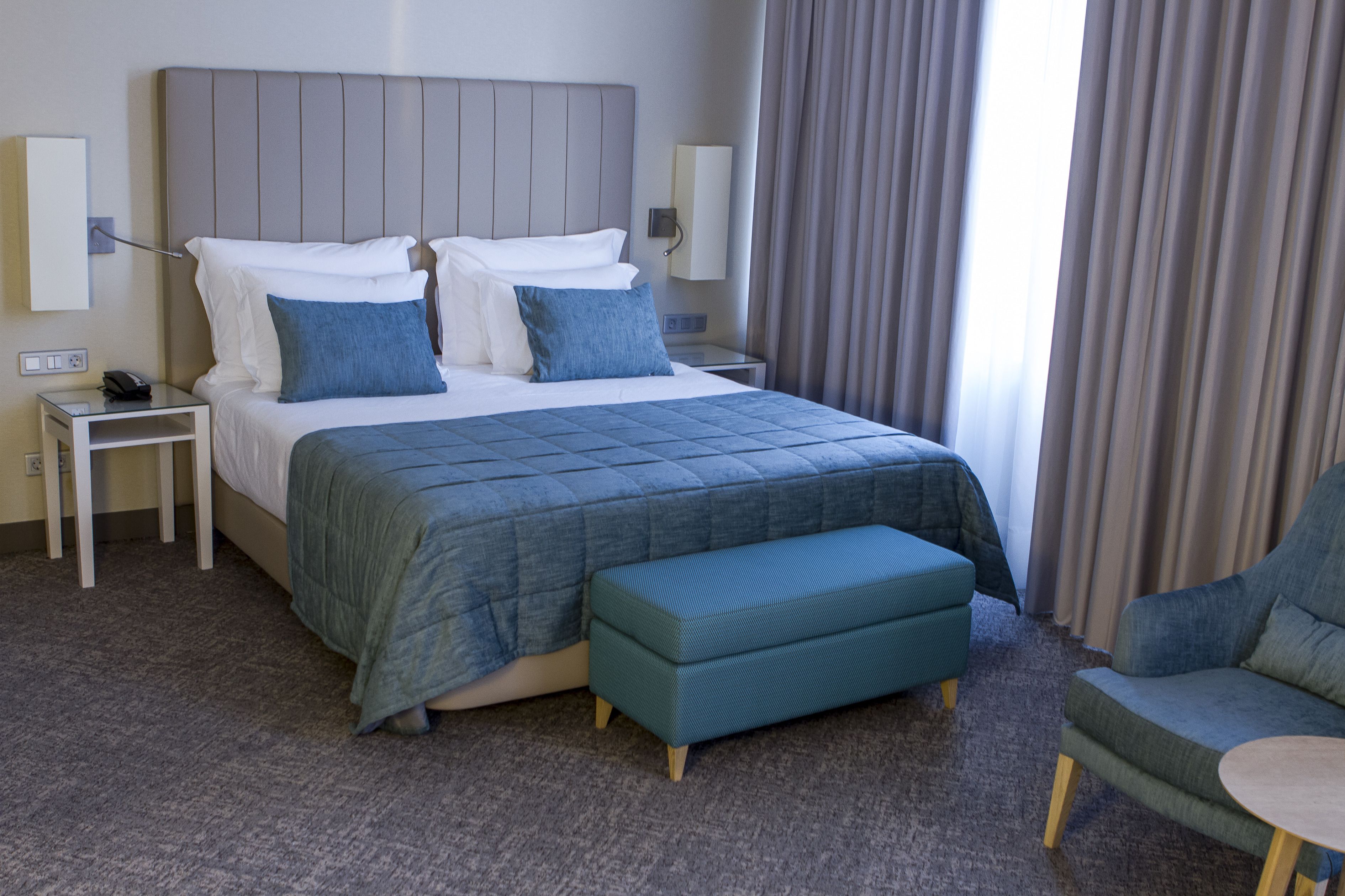 A hotel room with a large bed, a bedside table with a phone, a nightstand, a blue bench, and a chair.