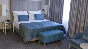 A hotel room with a large bed, a bedside table with a phone, a nightstand, a blue bench, and a chair.