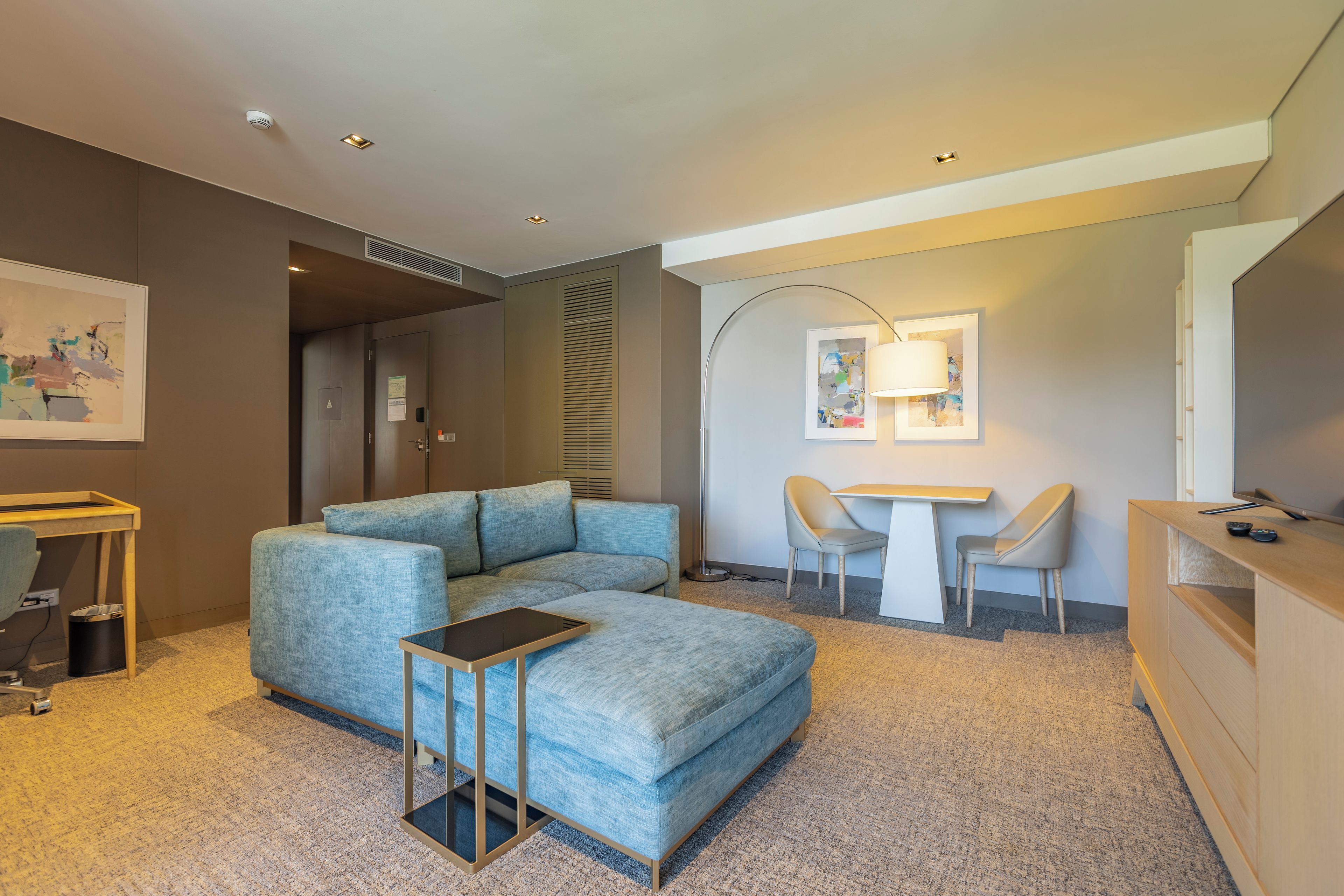 A modern, well-lit room with a sectional sofa, a small table, two chairs, a desk, and a TV.