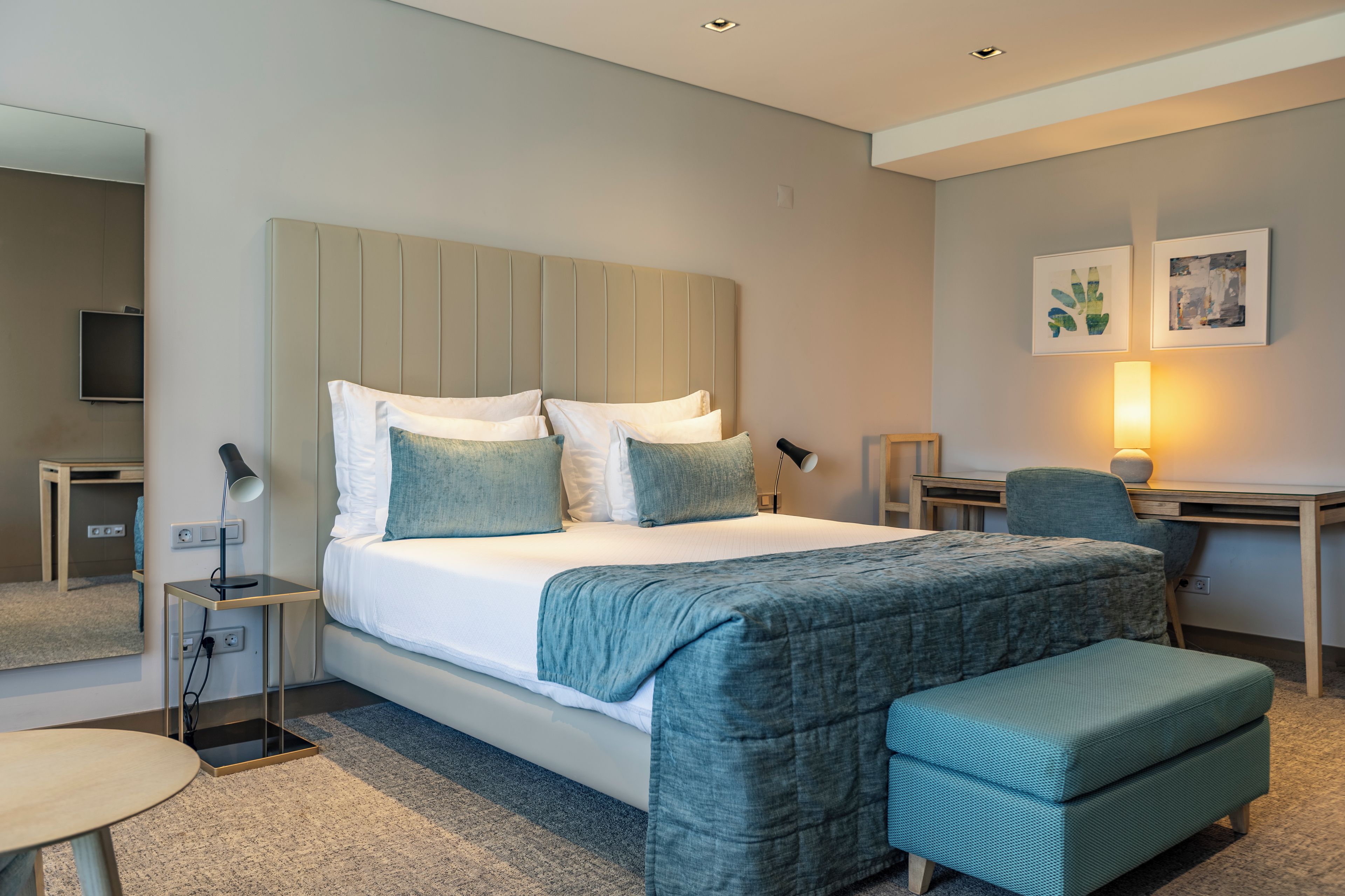 A modern hotel room with a large bed, bedside tables, a desk, a chair, and a small round table.