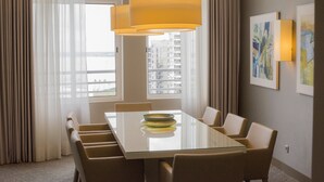Comedor de hotel con una mesa de vidrio, sillas color beige, una lámpara colgante amarilla y vistas a edificios a través de amplios ventanales.