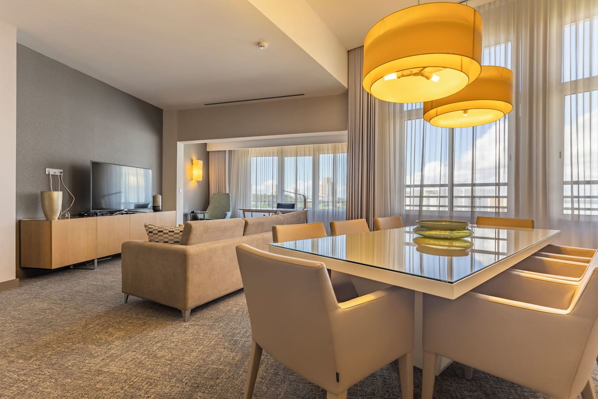 Une chambre d’hôtel moderne avec une table à manger en verre, un canapé beige et un téléviseur posé sur un meuble en bois.