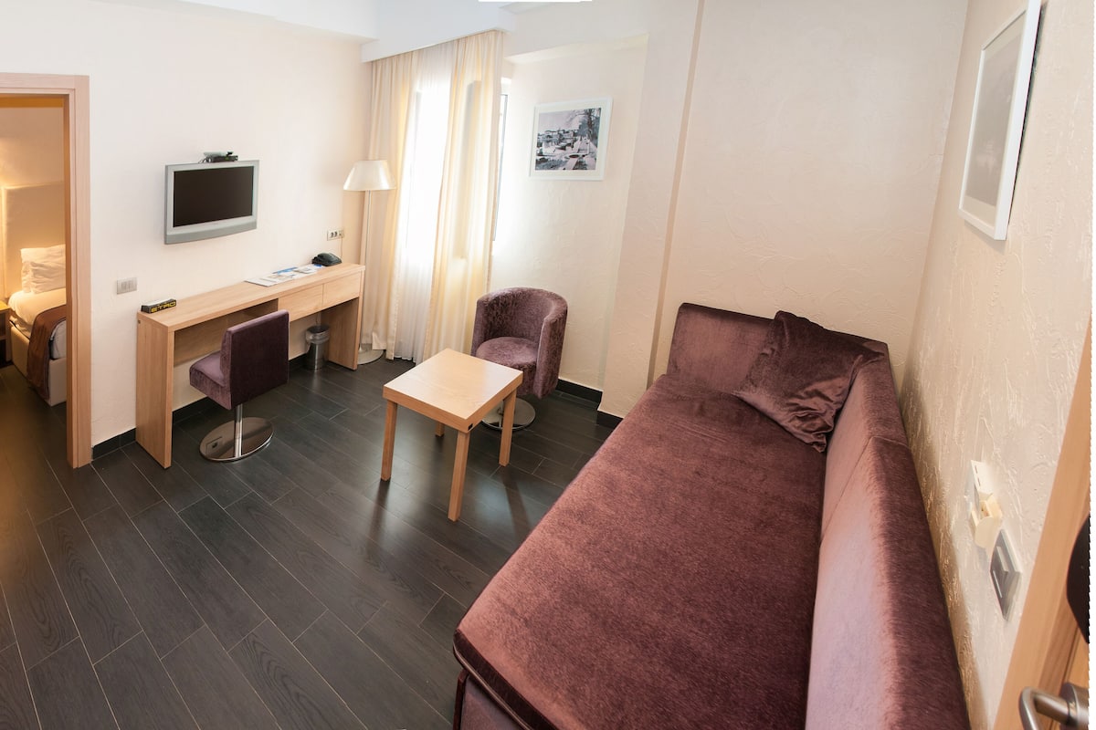 Ein gemütliches Hotelzimmer mit einem Holztisch, einem kleinen Tisch, einem violetten Sessel, einem braunen Sofa und einem an der Wand montierten Fernseher.