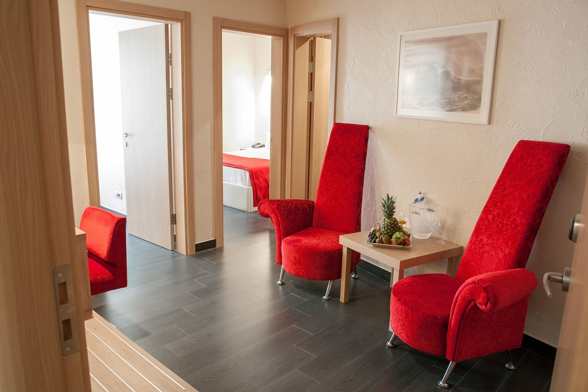 Ein Hotelzimmer mit Holzboden, roten Polsterstühlen, einem kleinen Tisch mit einer Ananas und einer Flasche sowie einem gerahmten Bild an der Wand.