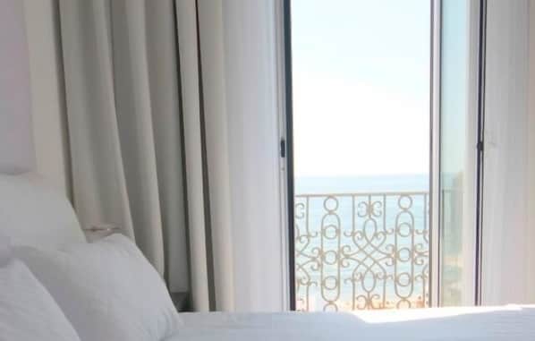 A hotel room with a large bed, a view of the sea through a balcony with decorative iron railings.