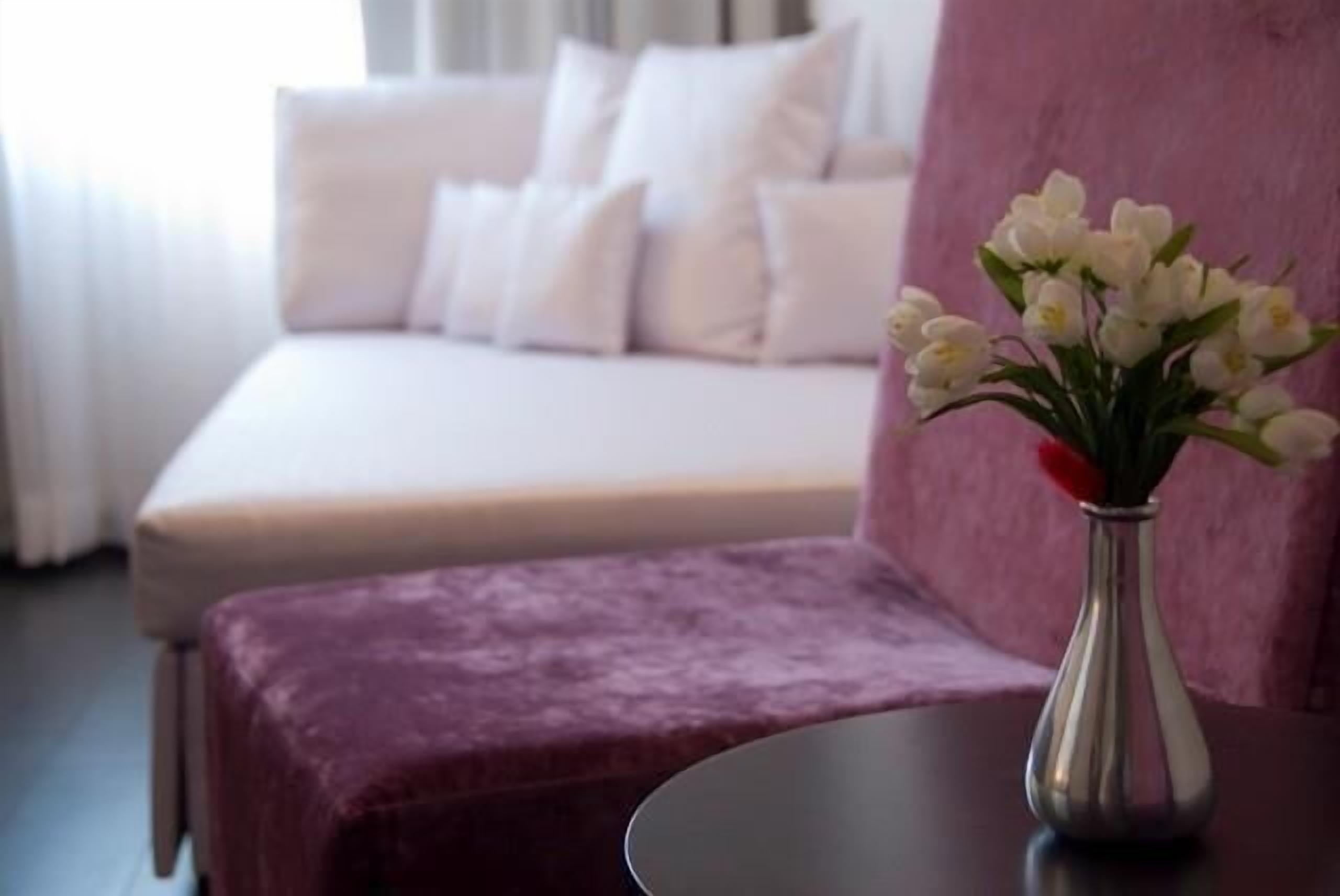 Une chambre à coucher avec un lit, une table de chevet ornée d’un vase de fleurs et un pouf violet.