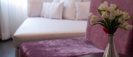 Une chambre à coucher avec un lit, une table de chevet ornée d’un vase de fleurs et un pouf violet.