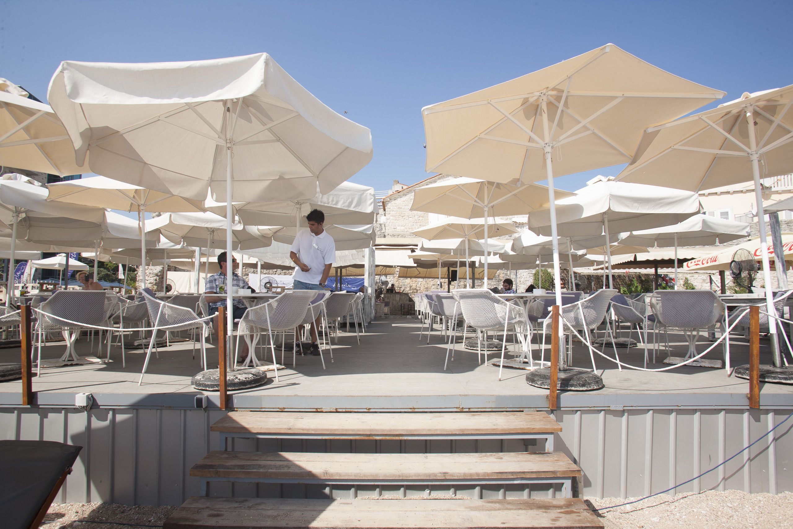 A terrace with multiple white umbrellas, wicker chairs, and a wooden staircase leading up.