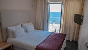 Ein Hotelzimmer mit Bett, Nachttisch aus Holz, Fernseher an der Wand und Meerblick durch das Fenster.