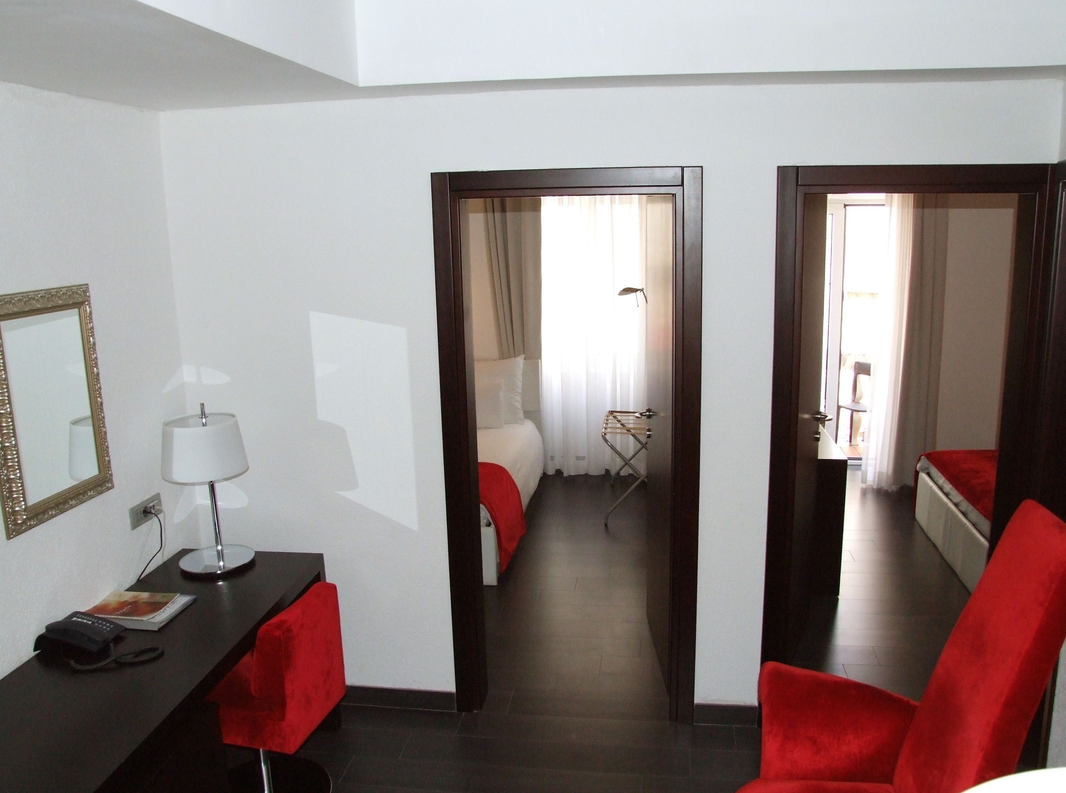 A hotel room with a desk, chair, and a mirror. There is a bed with a red blanket and a window with curtains.