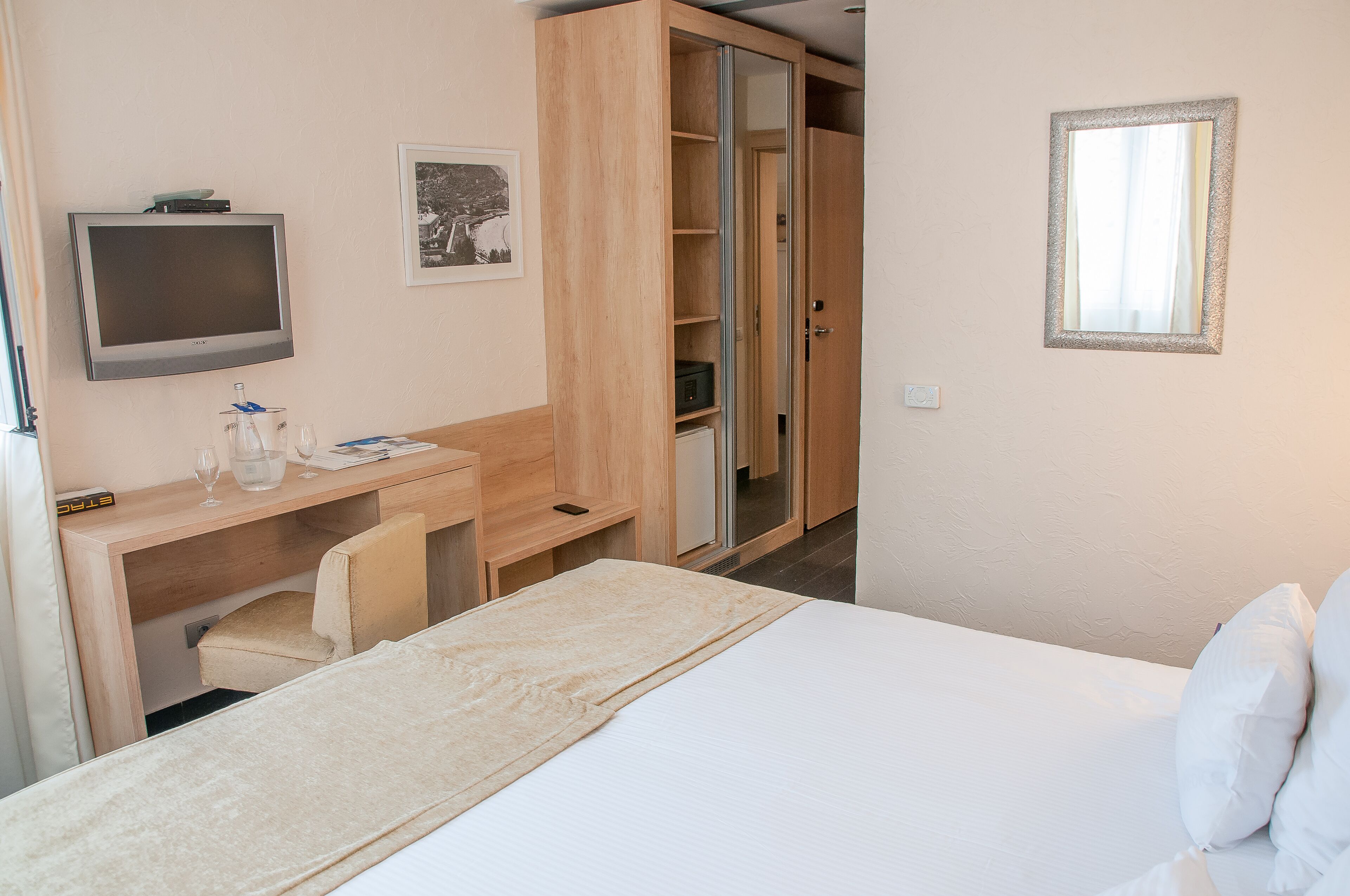 A hotel room with a bed, a desk with a chair, a television, a wardrobe, and a framed picture on the wall.