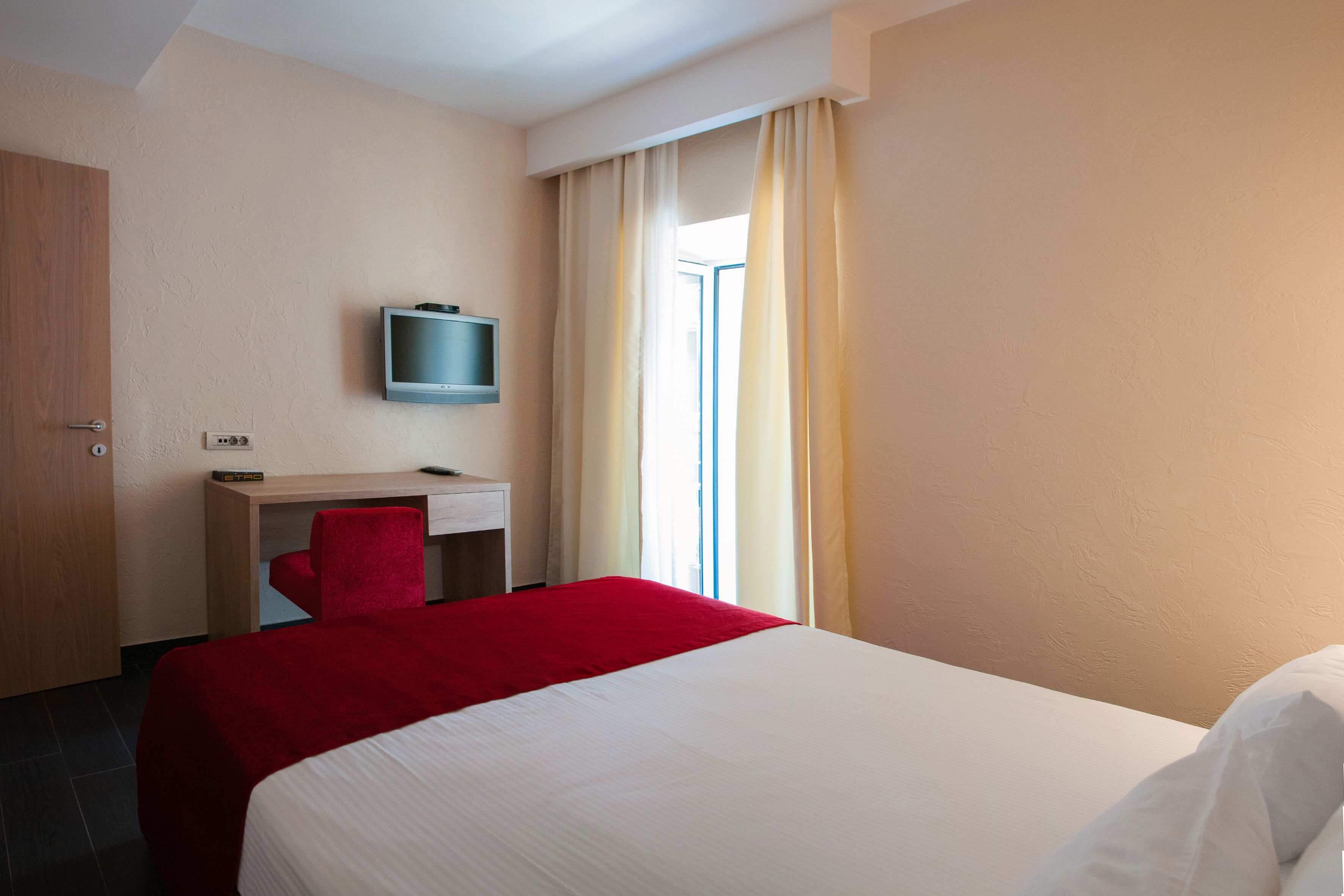 A hotel room with a bed, a desk with a chair, a television, and a window with curtains.
