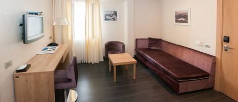 A compact living space with a wooden desk, a purple chair, a small table, a brown sofa, and a television mounted on the wall.