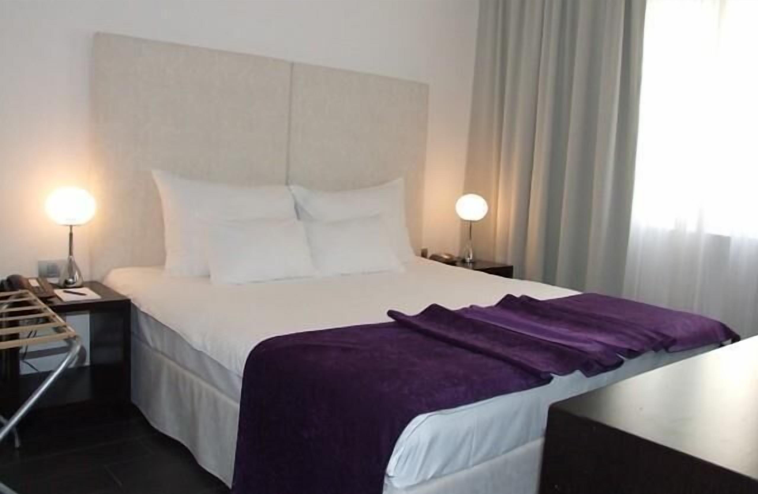 A hotel room with a bed, two bedside lamps, a nightstand, and a window with curtains.