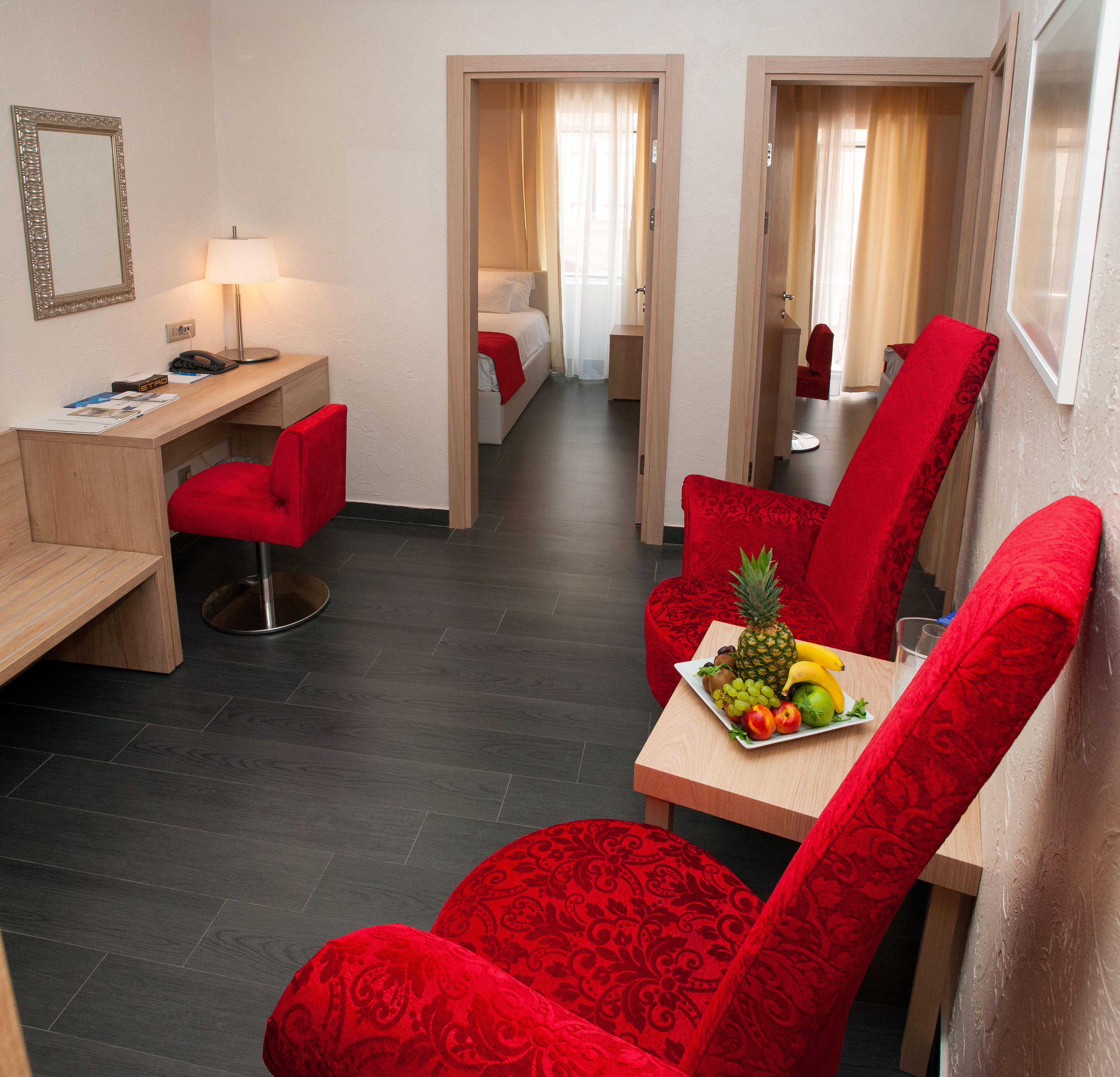 A hotel room with a desk, a red chair, a dining table with fruit, and a bed with curtains.