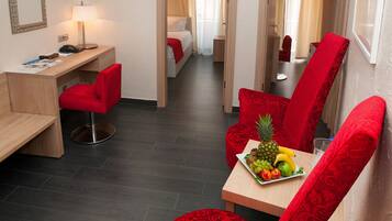 A hotel room with a desk, a red chair, a dining table with fruit, and a bed with curtains.