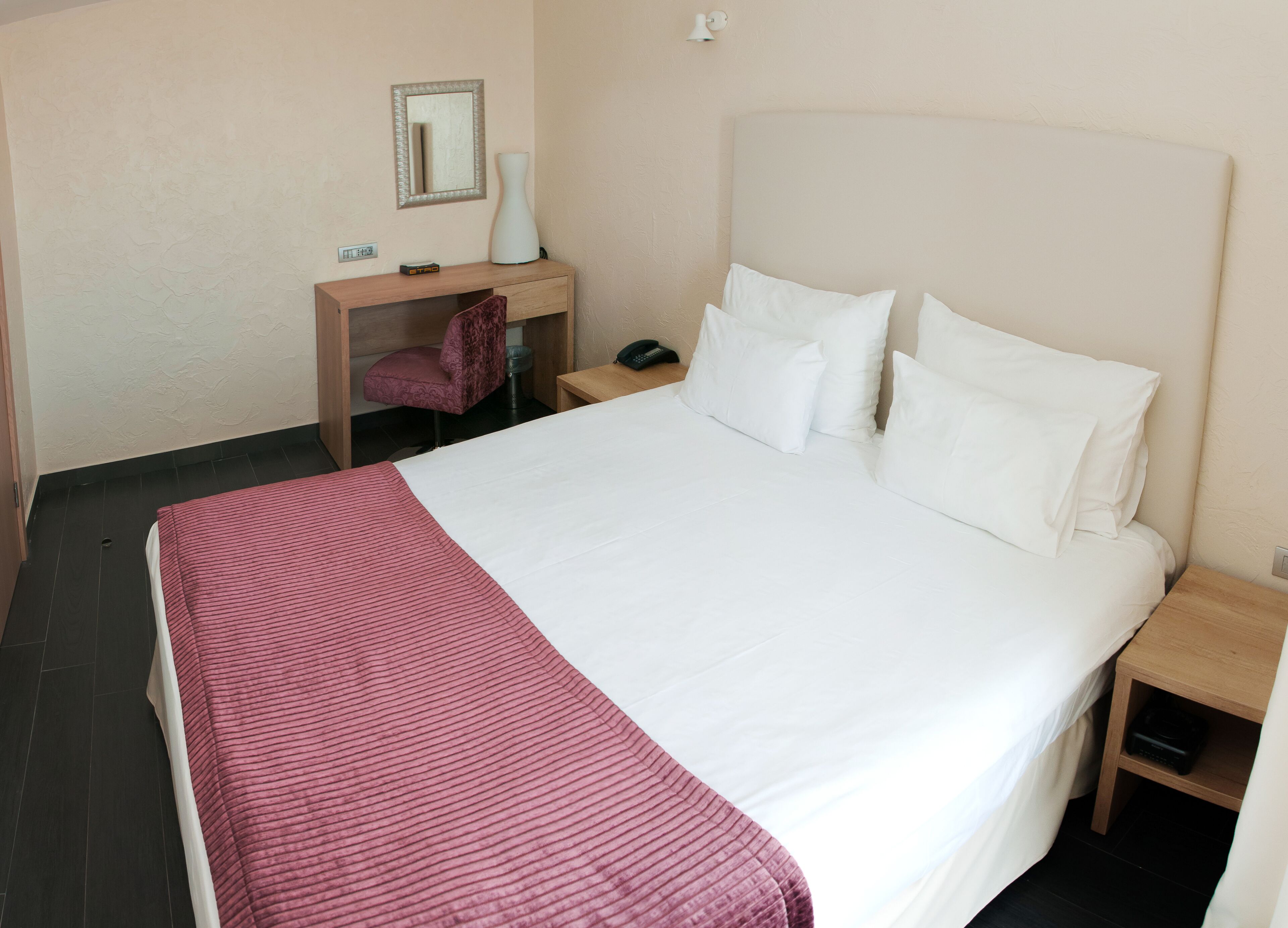 A hotel room with a large bed, a desk with a chair, a mirror, and a bedside table.
