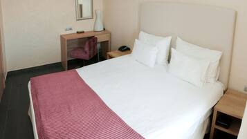 A hotel room with a large bed, a desk with a chair, a mirror, and a bedside table.