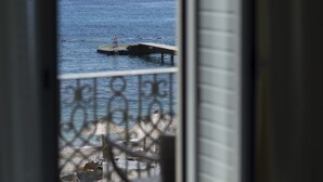 Blick auf einen Steg, der sich in ein ruhiges Meer erstreckt, mit einer kleinen Statue auf dem Steg und einem Strandbereich mit Sonnenschirmen im Vordergrund.