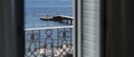A view of a pier extending into a calm sea, with a small statue on the pier and a beach area with umbrellas in the foreground.