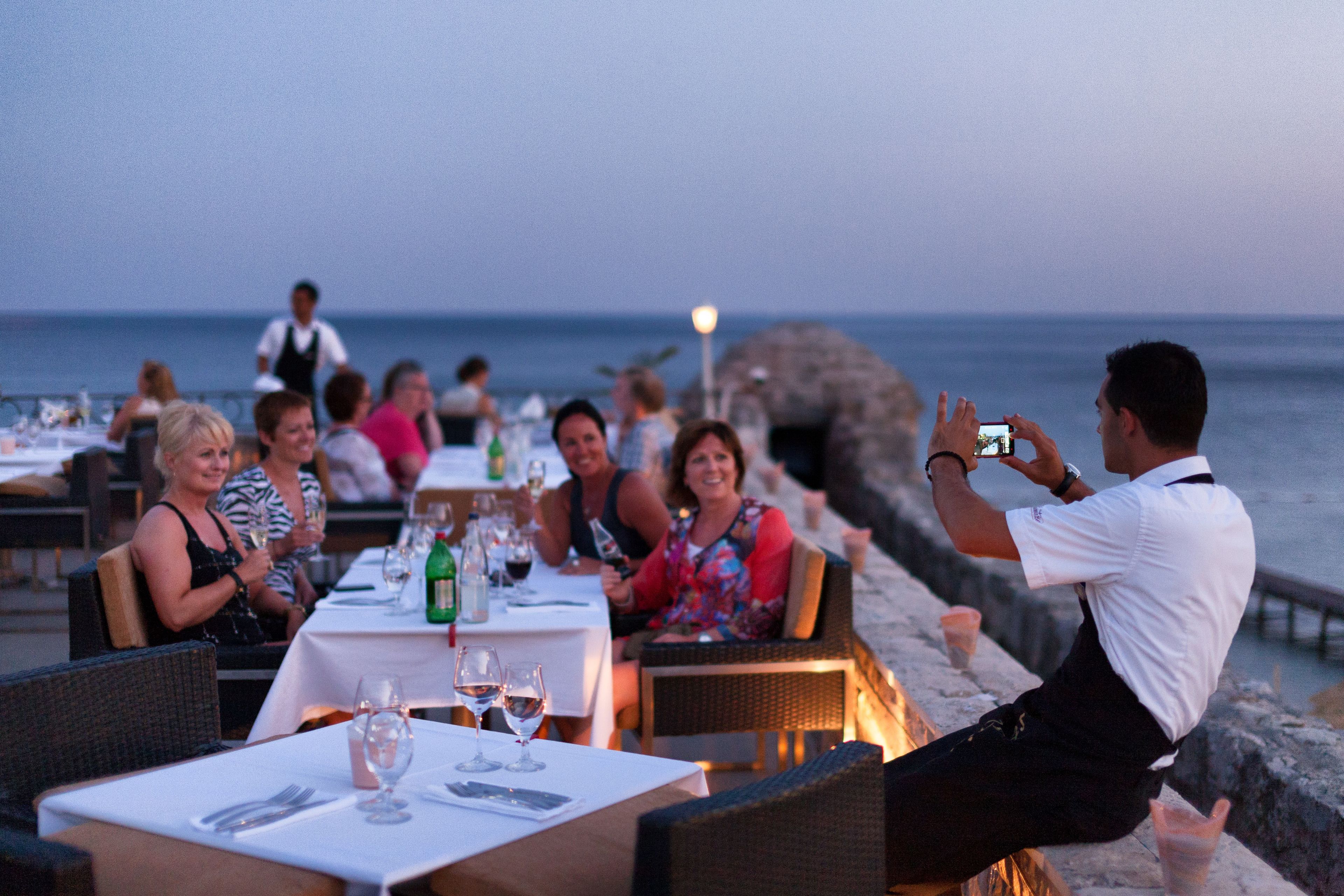 A restaurant with outdoor seating overlooking the sea, tables set with wine glasses and bottles, and a person taking a photo.