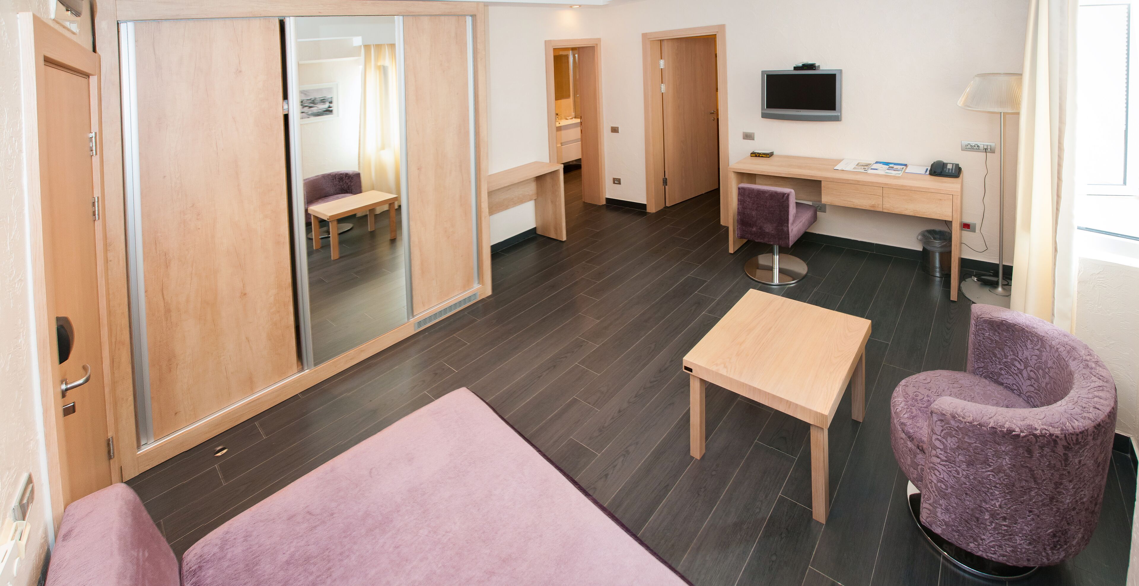 Una camera d'albergo moderna con una scrivania in legno, una sedia viola, un tavolino e uno specchio grande.