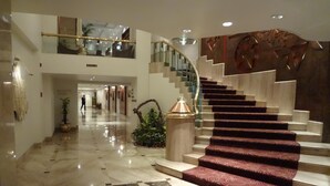 Een ruime lobby met een marmeren vloer, een imposante trap met rode lopers en een glazen balustrade.