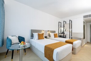 Hotellihuone, jossa on kaksi sänkyä, pieni pöytä, jossa on hedelmiä, sininen nojatuoli ja peili.