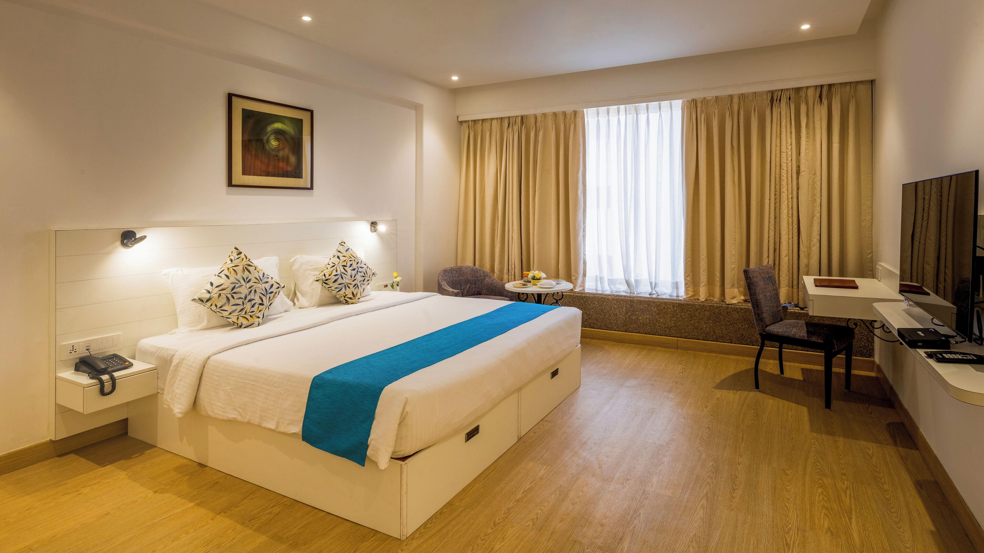 A hotel room with a large bed, bedside table with a phone, a desk with a chair, a TV, and a window with curtains.