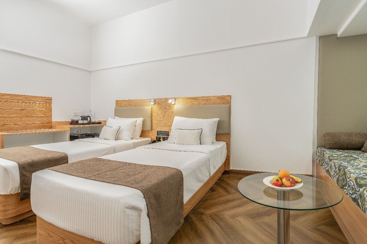 A hotel room with two beds, a small table with fruit, and a separate seating area with a patterned sofa.