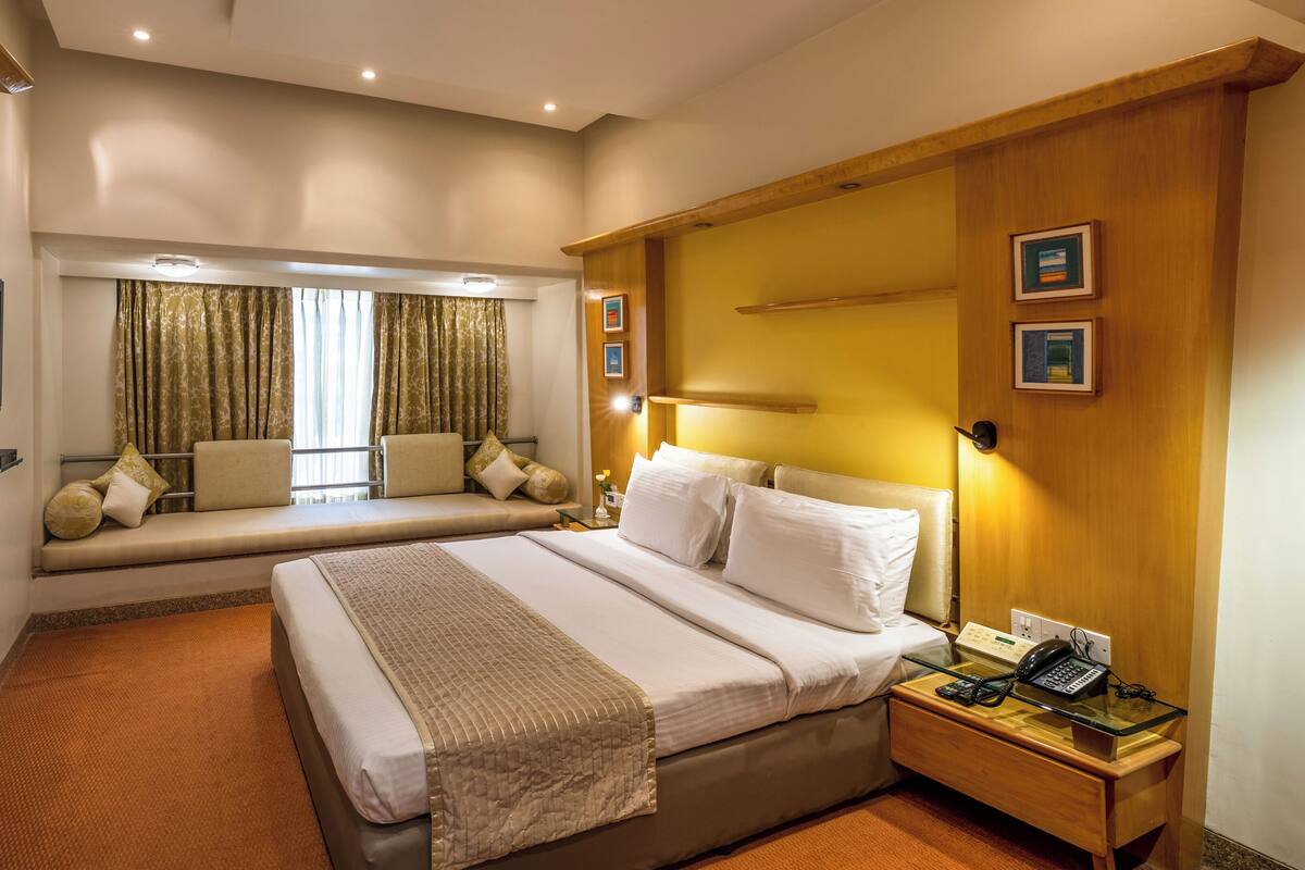 Una habitación de hotel con una cama grande, una mesita de noche con teléfono, un televisor montado en la pared y una ventana con cortinas.