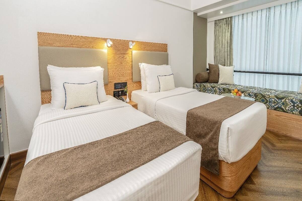 Habitación de hotel con dos camas, cabecero de madera, mesitas de noche, una mesita pequeña con un frutero y una ventana con cortinas.