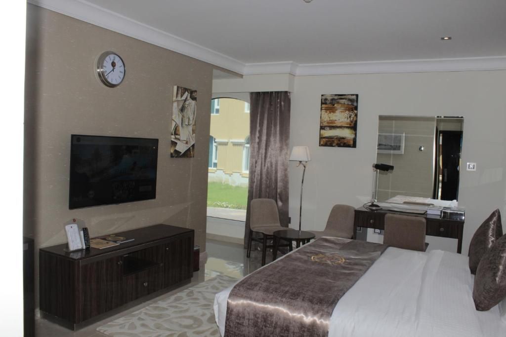 Habitación de hotel con cama, escritorio, silla, televisor, reloj y ventana con cortinas.