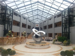 Un amplio atrio con una escultura central, techo de cristal y una zona de estar con sofá y sillones.