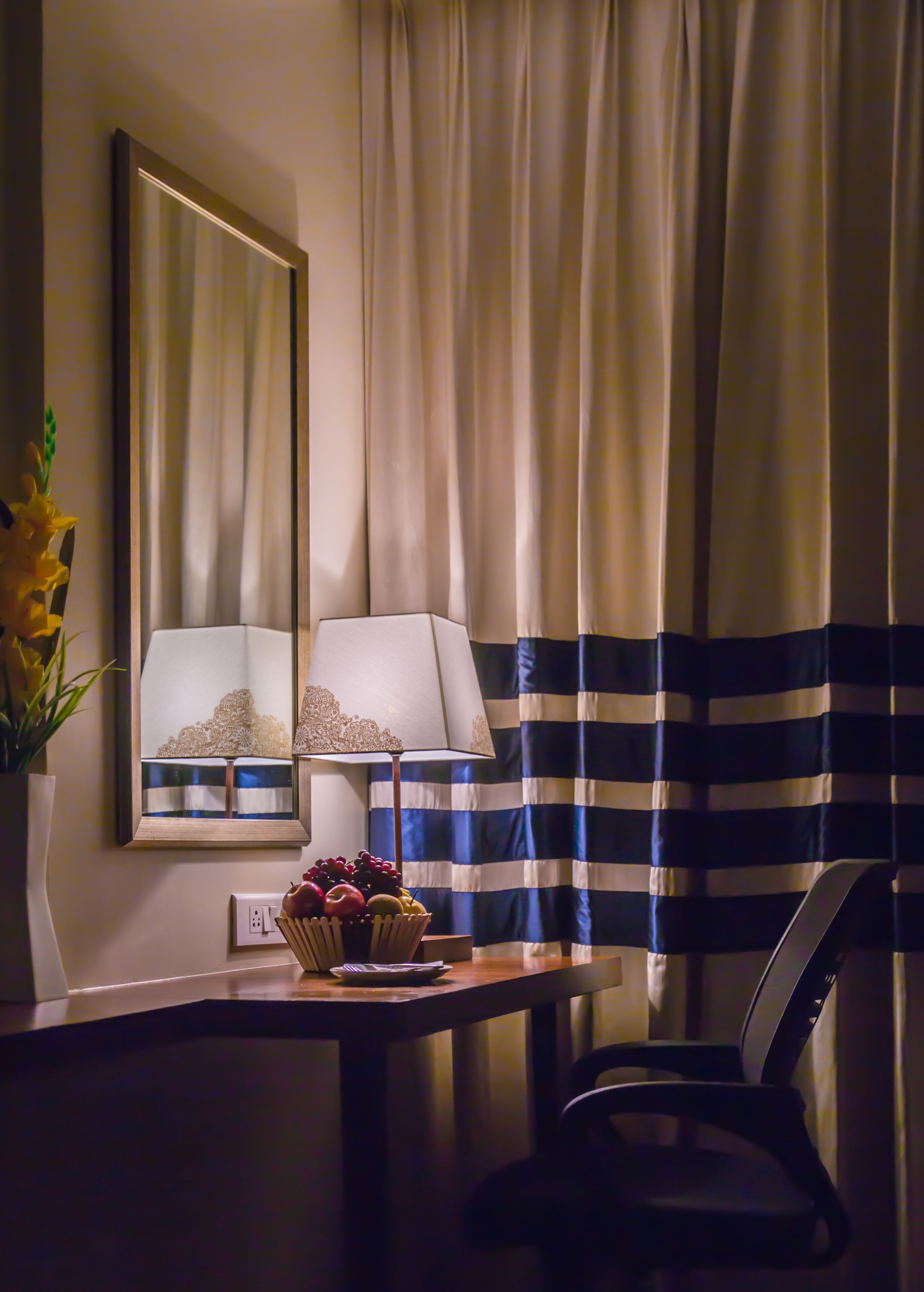 A hotel room with a desk, a chair, a lamp, a mirror, and a view of the outside through curtains.