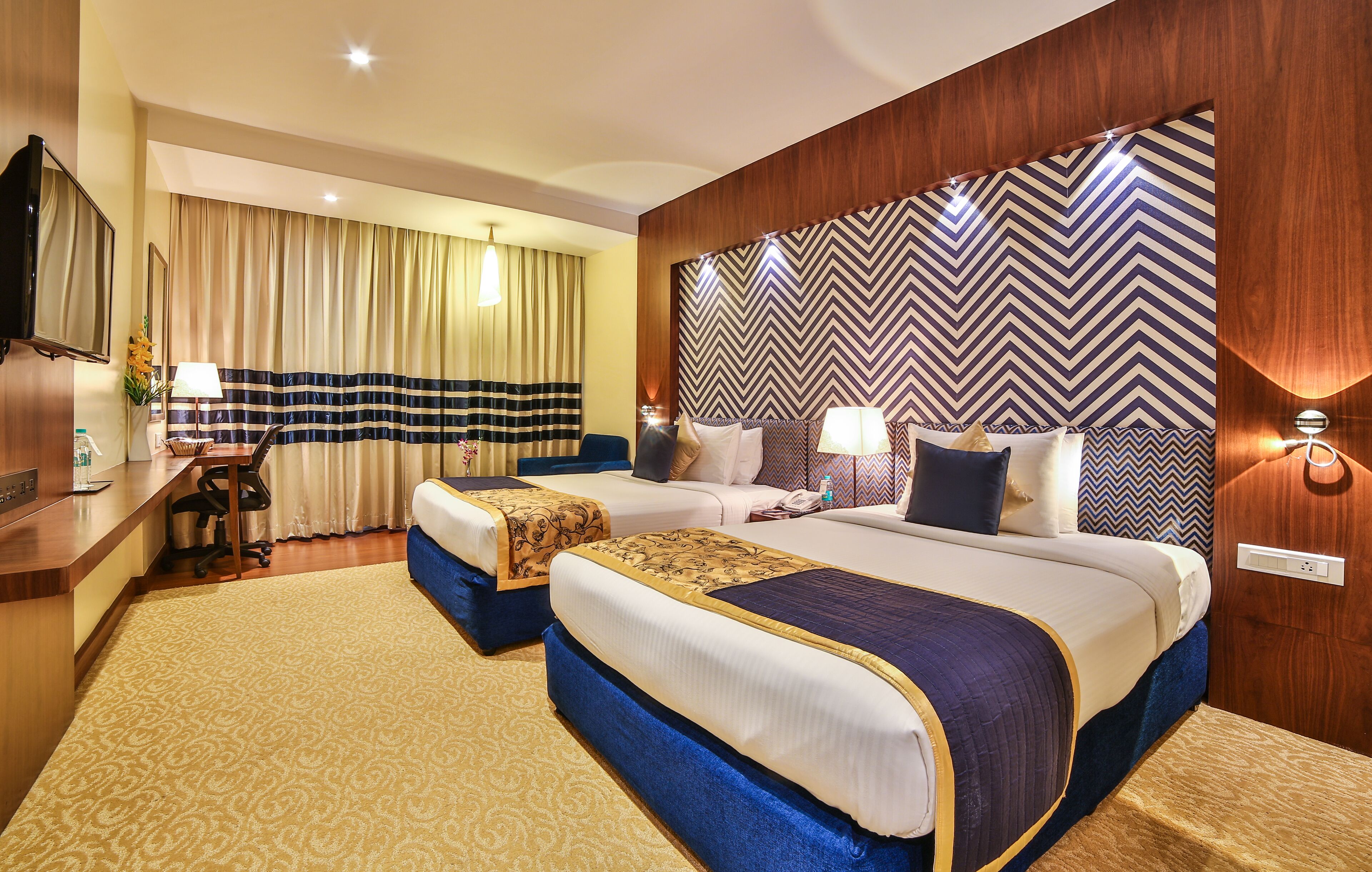 A hotel room with two beds, a desk with a chair, a TV, and a large mirror.